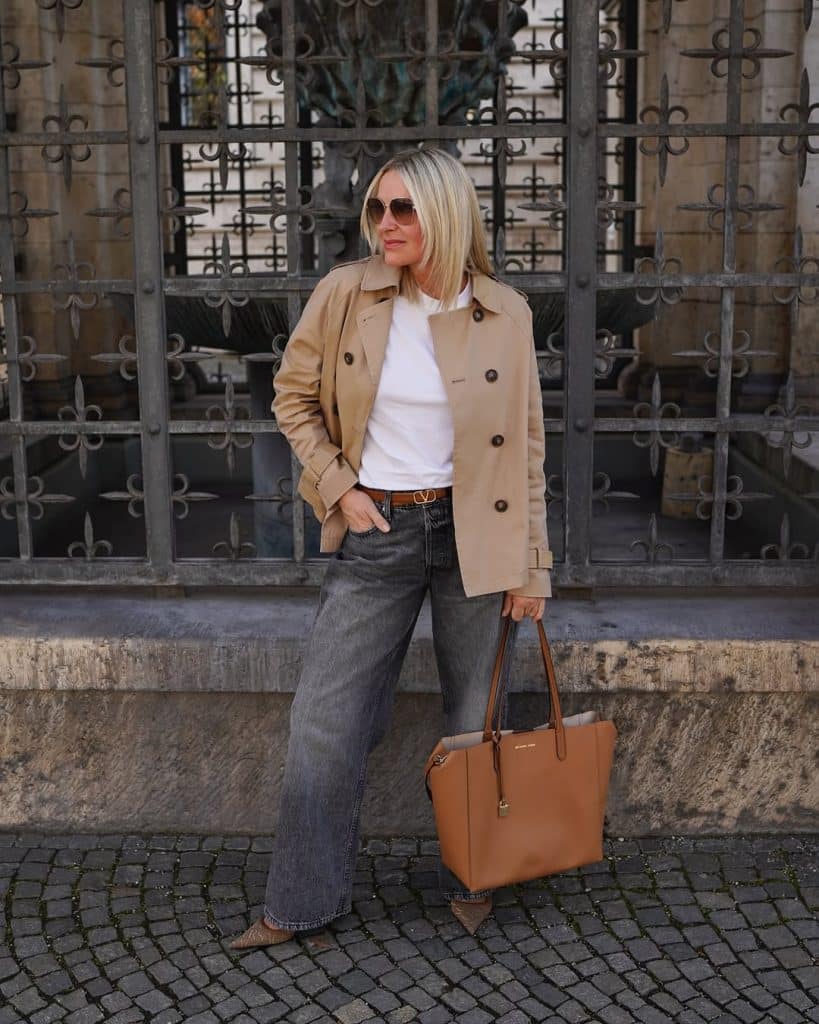 A blonde woman in a beige trench coat and wide-leg jeans posing on a cobblestone street.