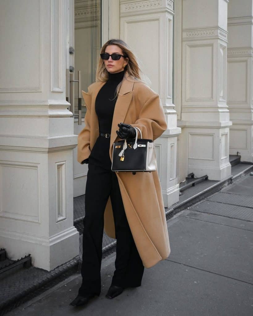 Woman in a tailored camel wool coat and black trousers carrying a luxury handbag.