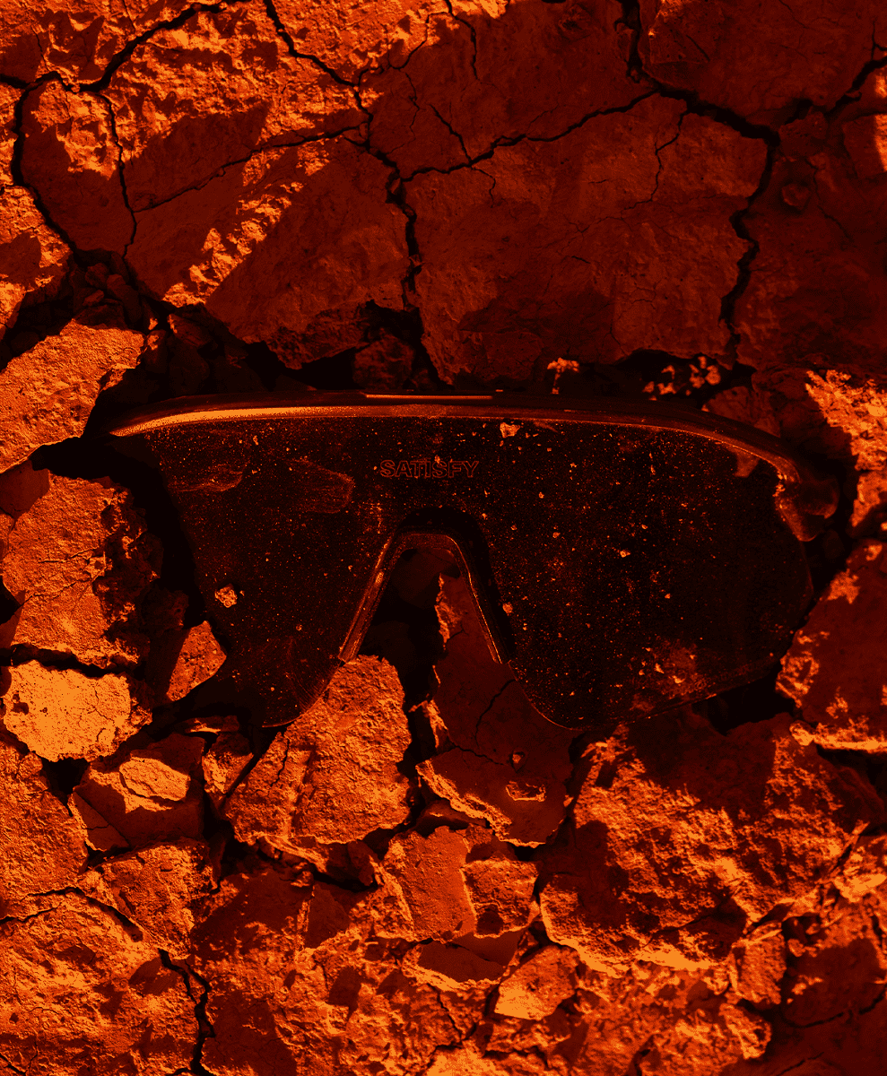 Satisfy brand goggles resting on dry, cracked desert earth under a warm orange glow.