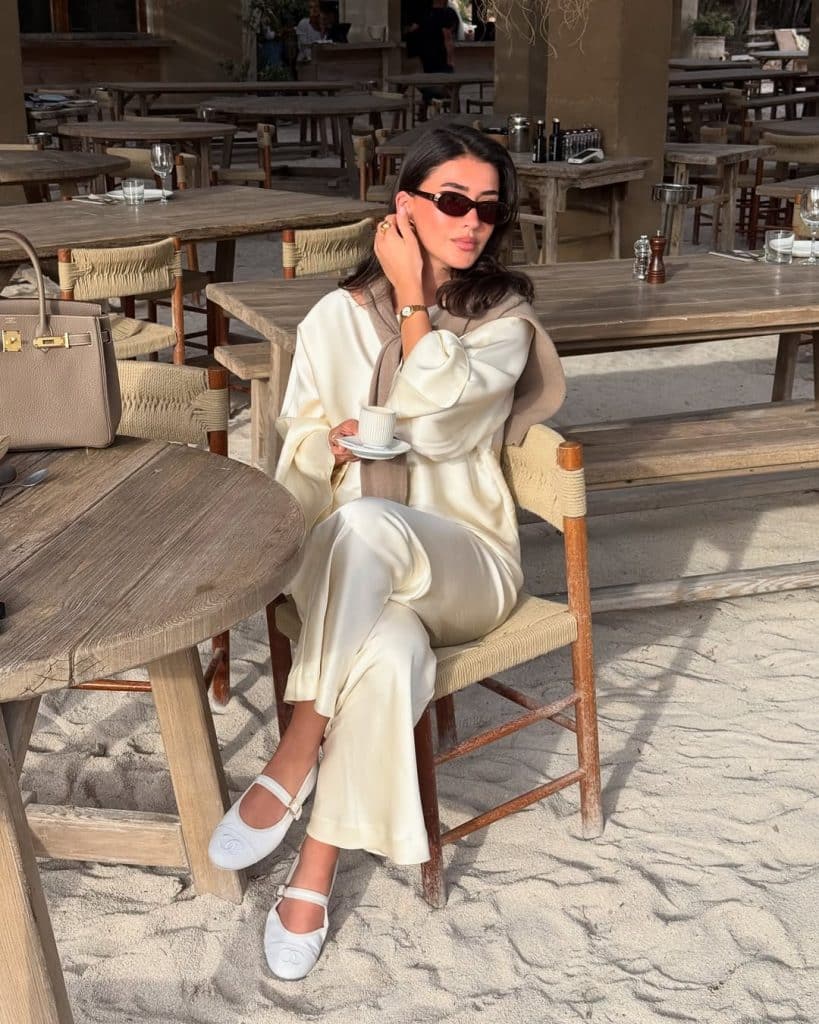 A woman in a creamy satin jumpsuit sitting at a sunlit beachside café.