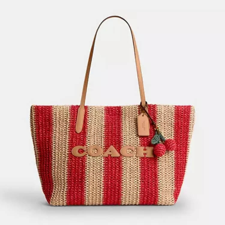 A Coach woven straw tote with red and beige stripes and a decorative cherry charm.