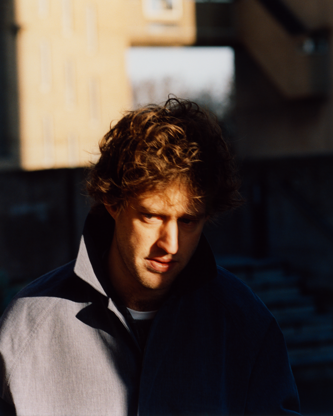 A man in a two-tone light gray and dark blue jacket posing in an urban, sunlit environment.