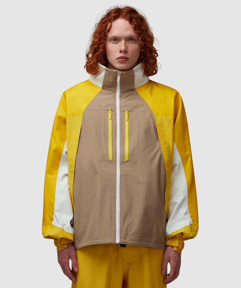 A model wearing a mustard yellow, beige, and white color-blocked technical jacket from the Nike Nocta collection.