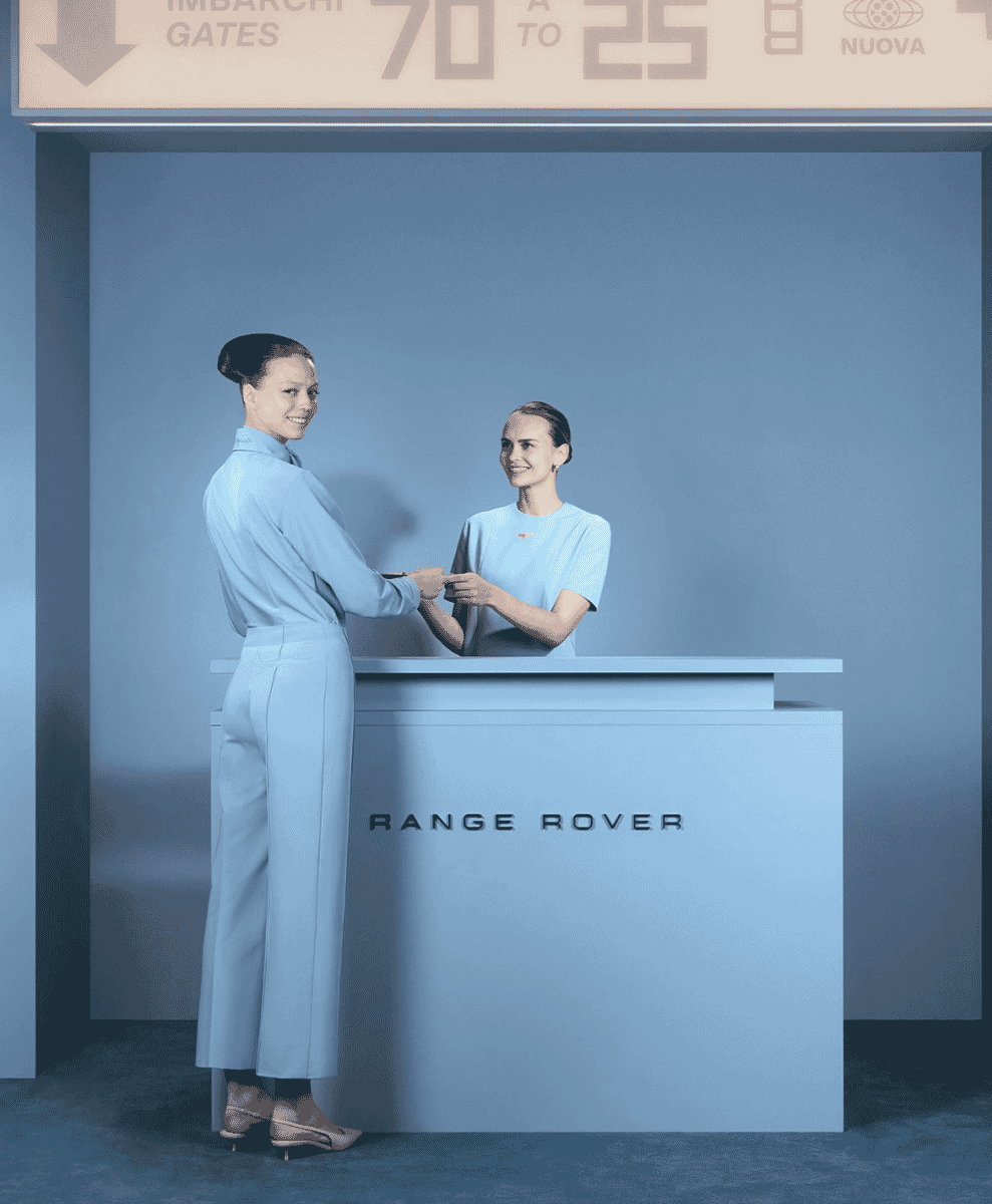 Two women in light blue professional wear at a minimalist Range Rover branding counter.