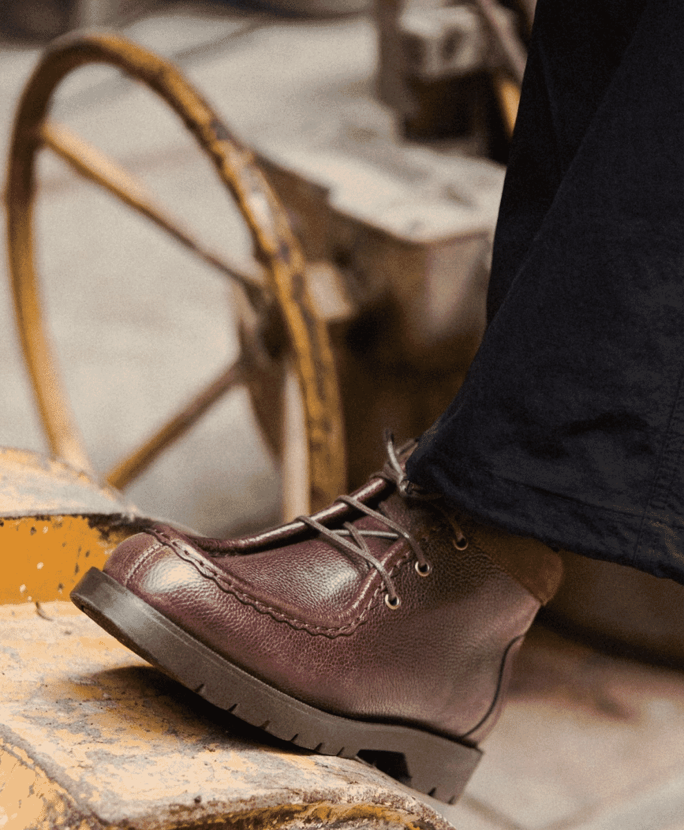 Brown leather lace-up boots standing next to a rusty wheel in an industrial setting.