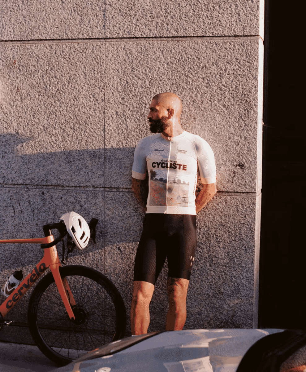 A man in a white cycling jersey standing beside an orange Cervelo bike against a textured wall.