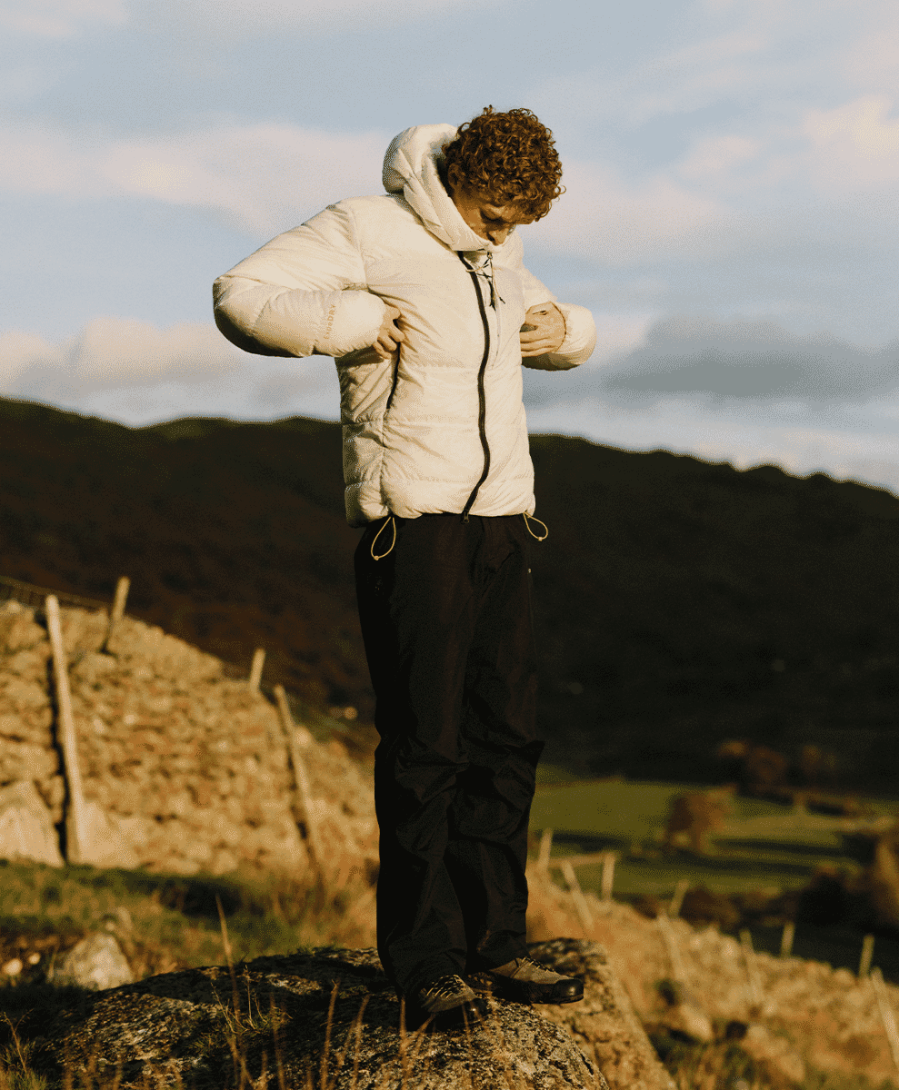 A man adjusting his white puffer jacket while standing on a hillside with rolling green mountains in the background.