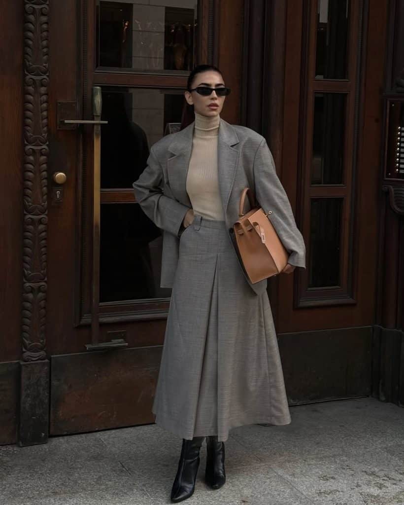 A woman wearing a grey oversized blazer and matching pleated midi skirt with black boots.