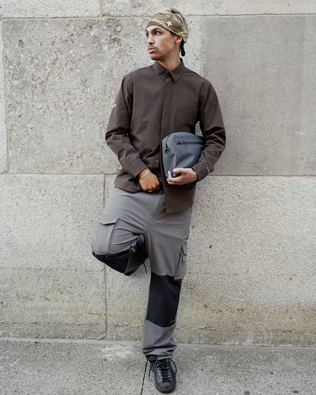 Man in a brown button-up shirt and camouflage beanie holding a grey tech pouch.