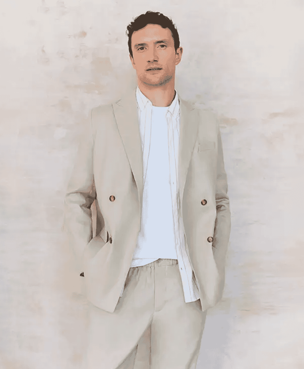 Man modeling a light beige double-breasted blazer against a textured off-white wall.