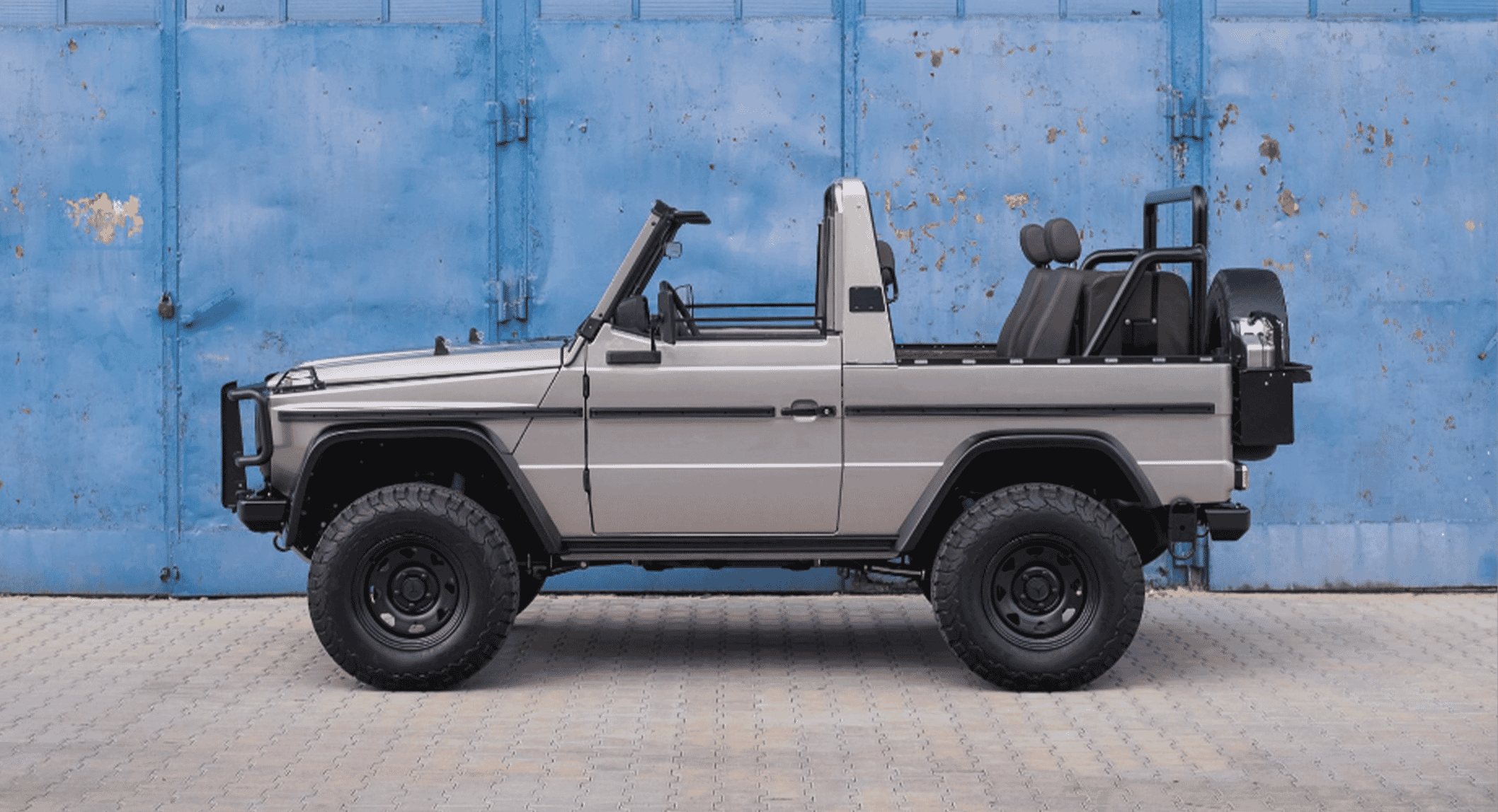 Full shot of a silver Mercedes 250 GD convertible pickup restoration against a blue industrial door.