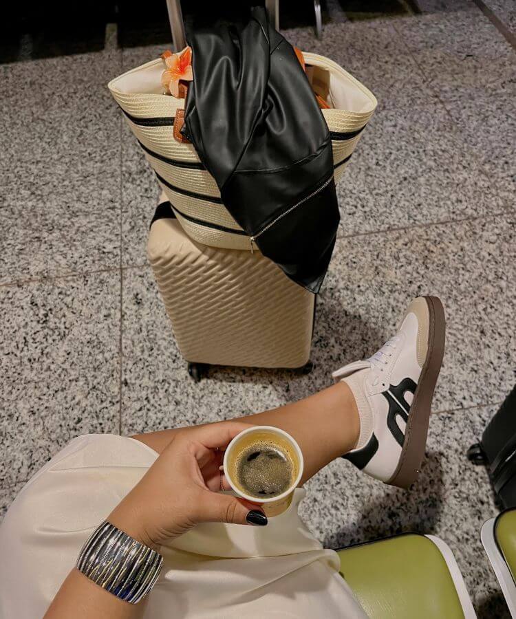 A person in a white dress and sneakers sits in an airport terminal holding a coffee cup with a suitcase nearby.
