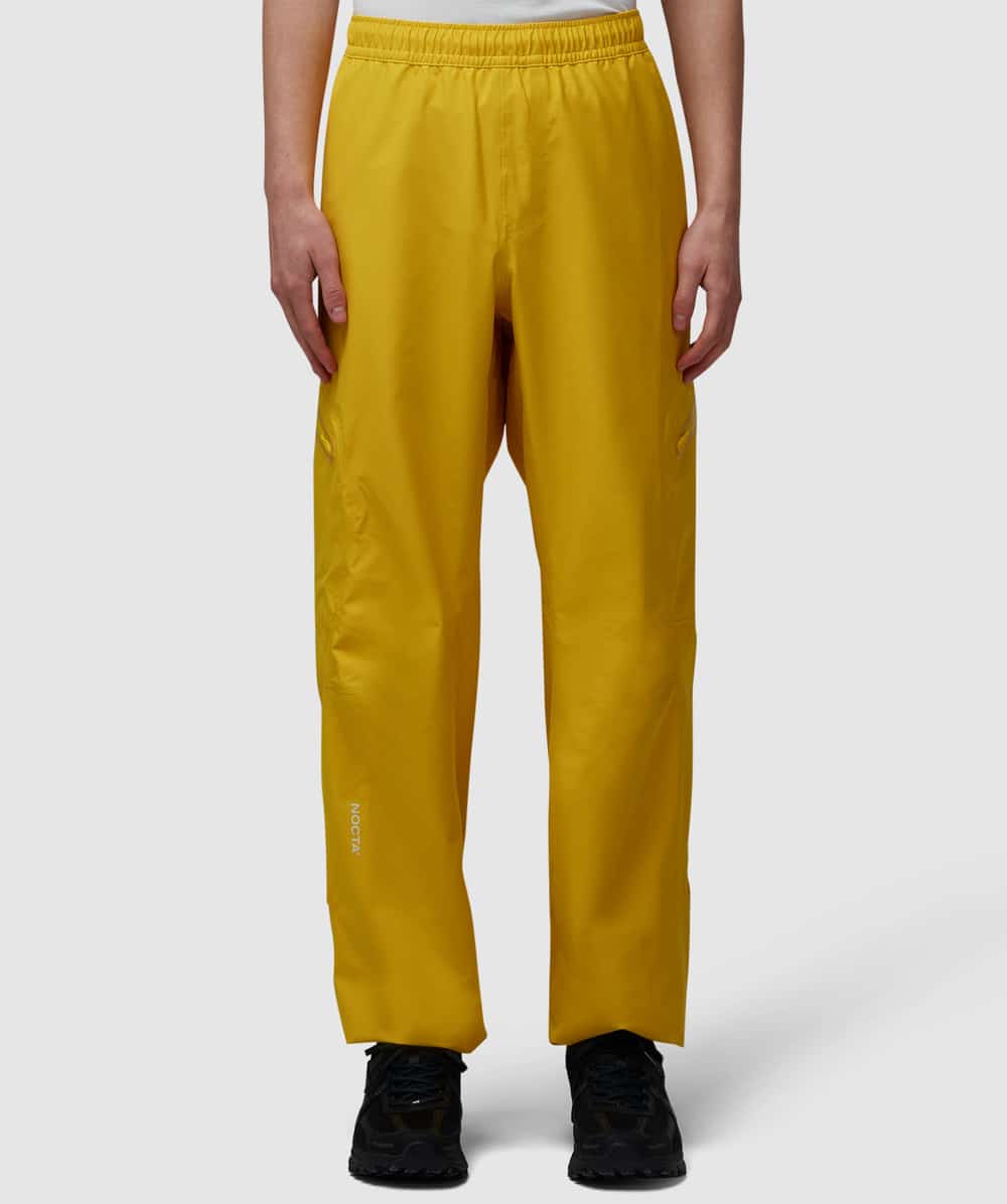 Model wearing mustard-yellow cargo-style pants with side pockets and elastic waist, paired with black technical sneakers.