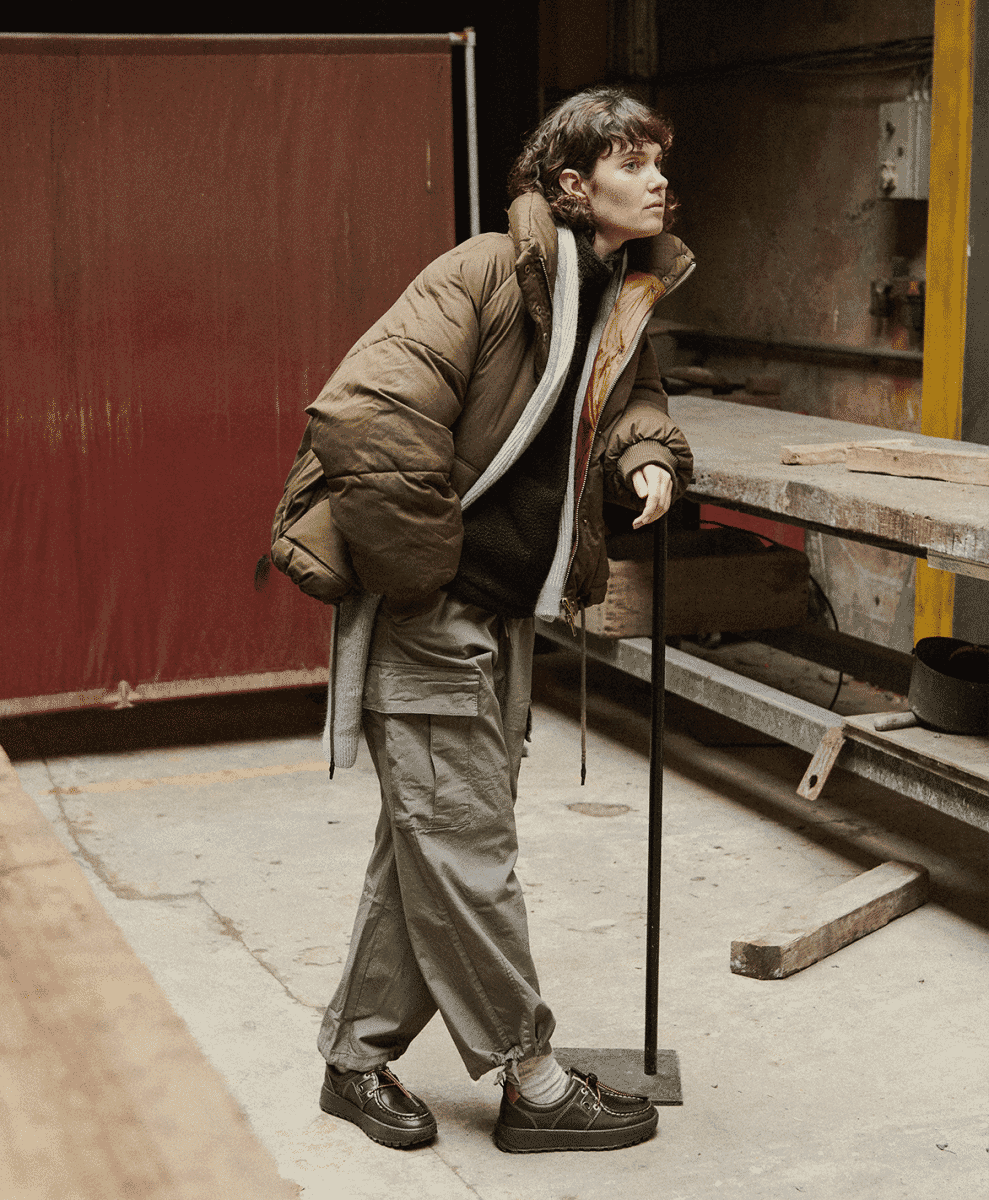 Person wearing a voluminous brown puffer jacket and cargo pants in an industrial setting.