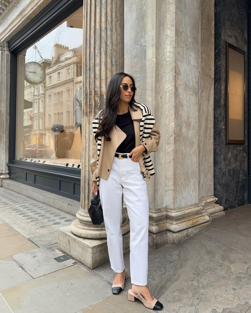A woman in a beige trench coat and white trousers posing against a marble column on a city street.