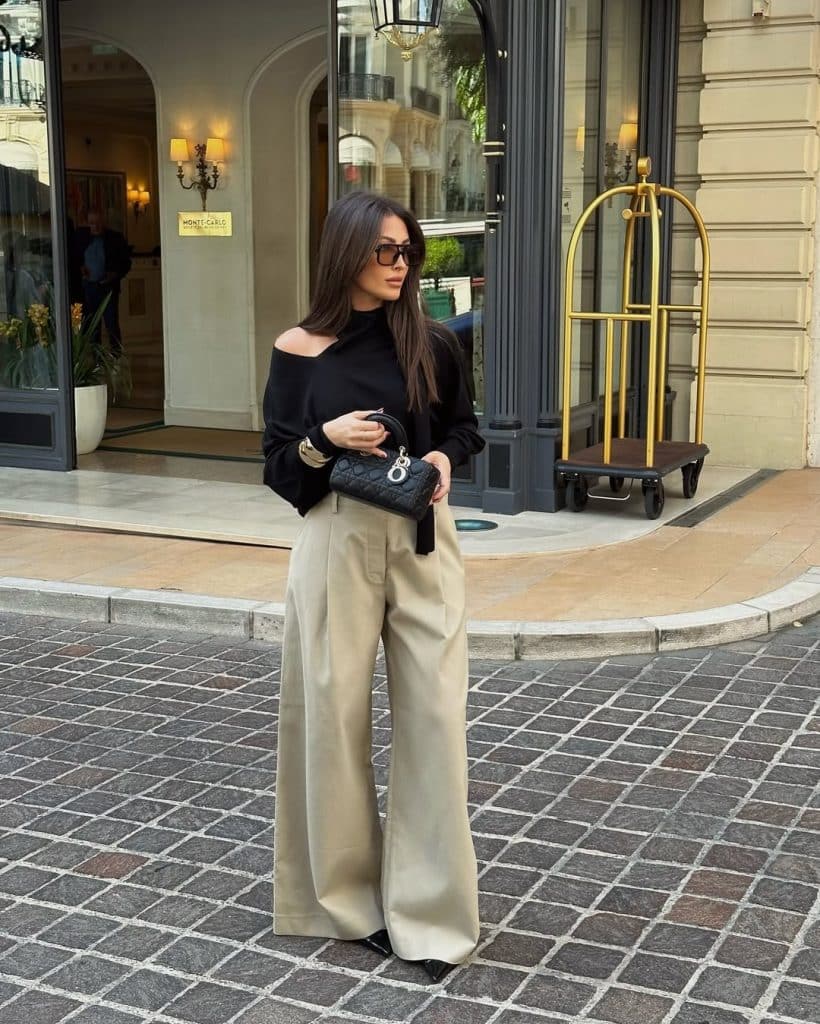 A woman wearing a black off-the-shoulder top with wide-leg beige trousers on a cobblestone street.