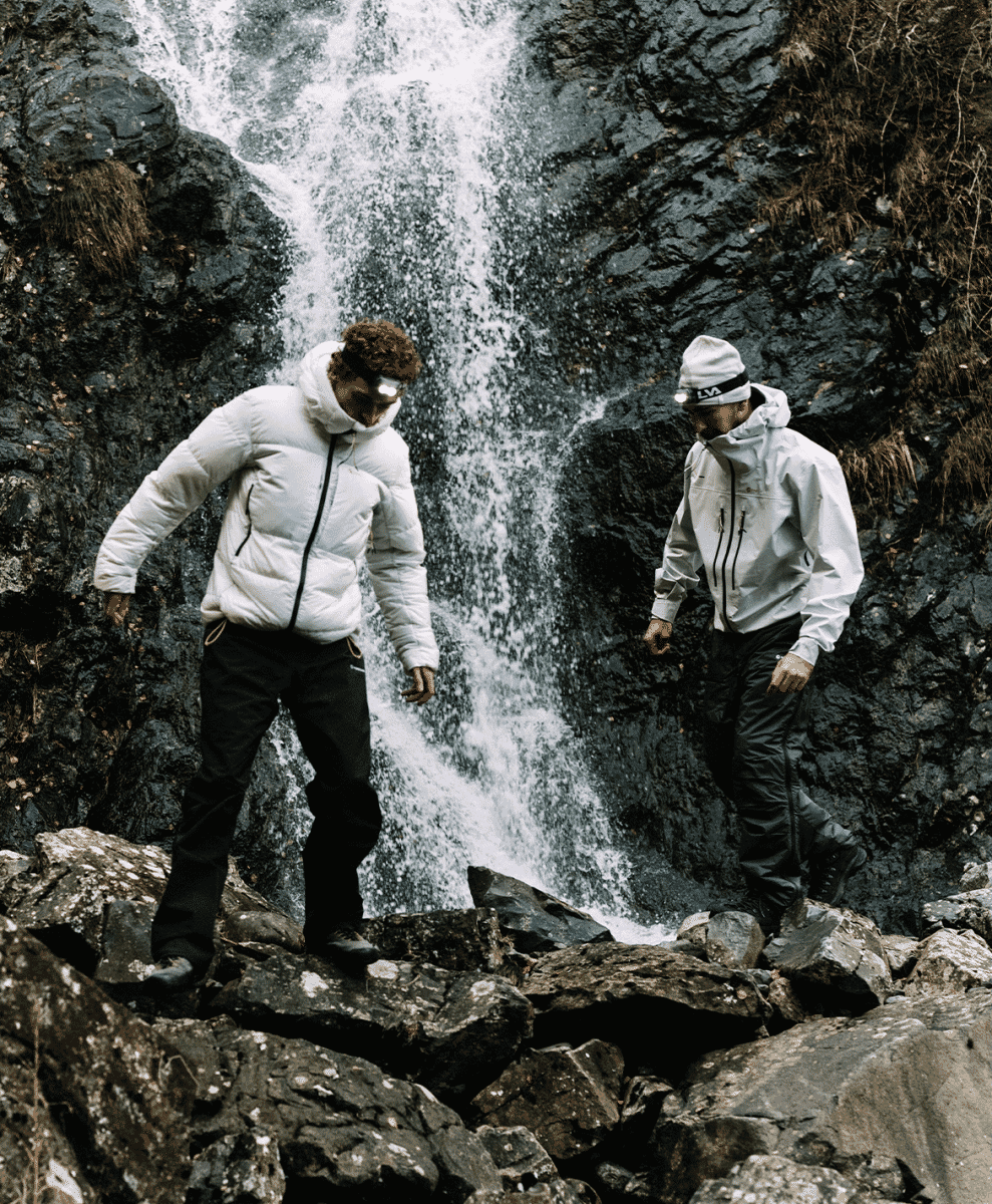 Two hikers in white insulated gear and headlamps standing by a waterfall in dark, rocky conditions.