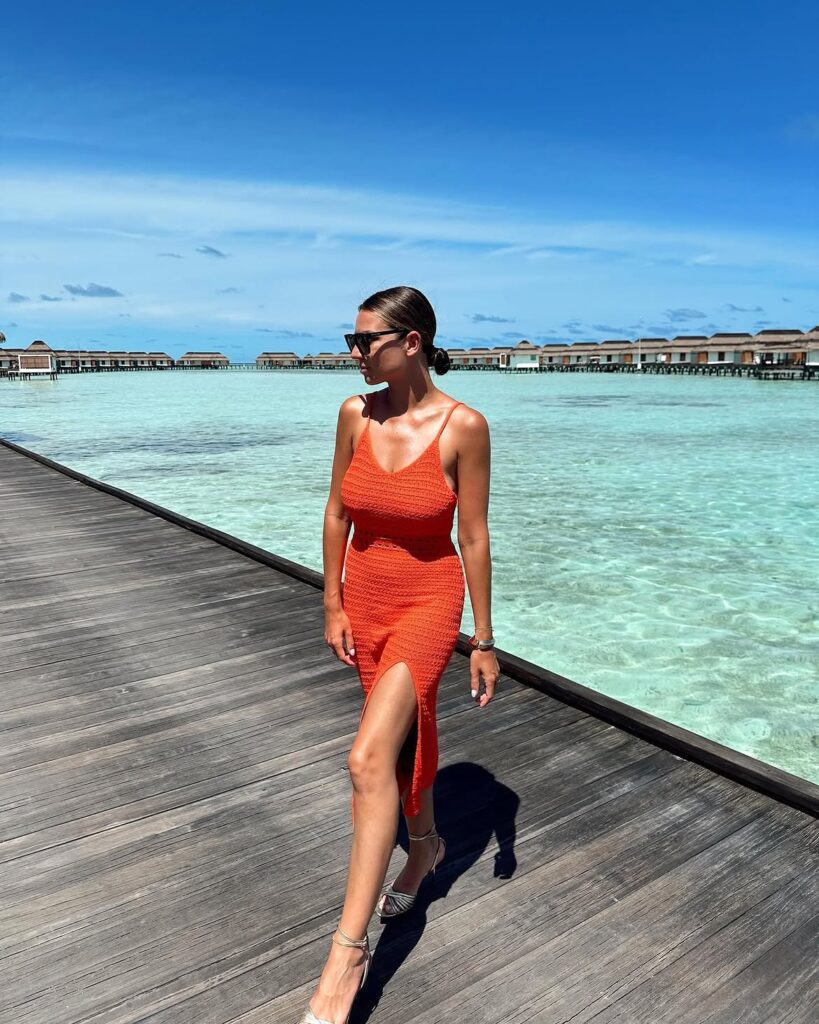 A woman in a vibrant orange textured knit midi dress walking along a pier with overwater bungalows behind her.