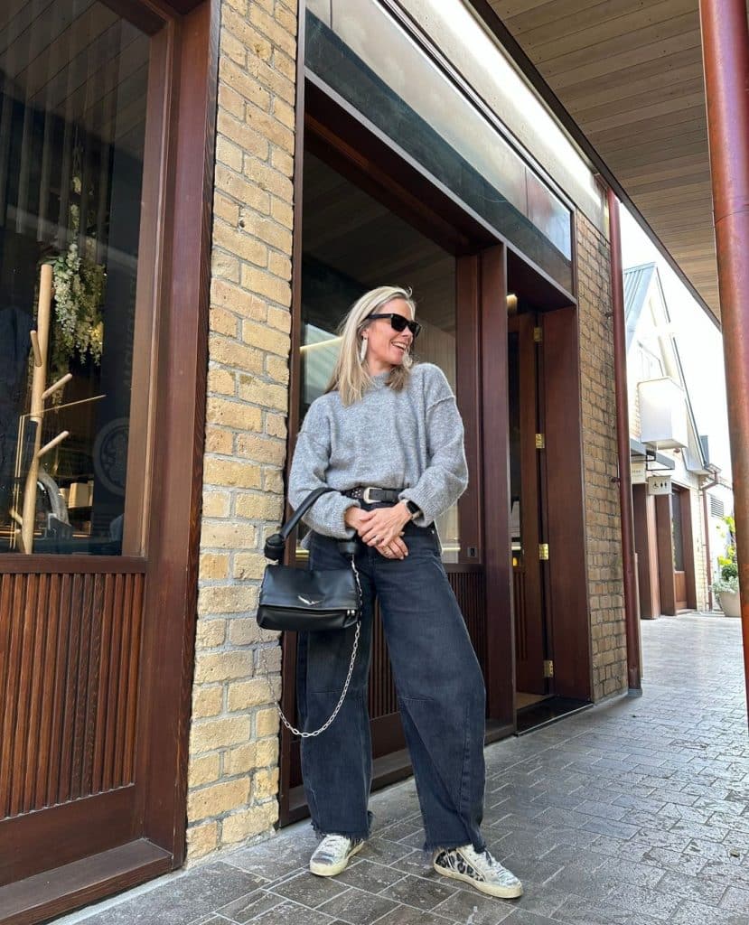 Woman in a grey oversized sweater and dark barrel jeans with patterned sneakers on a brick street.