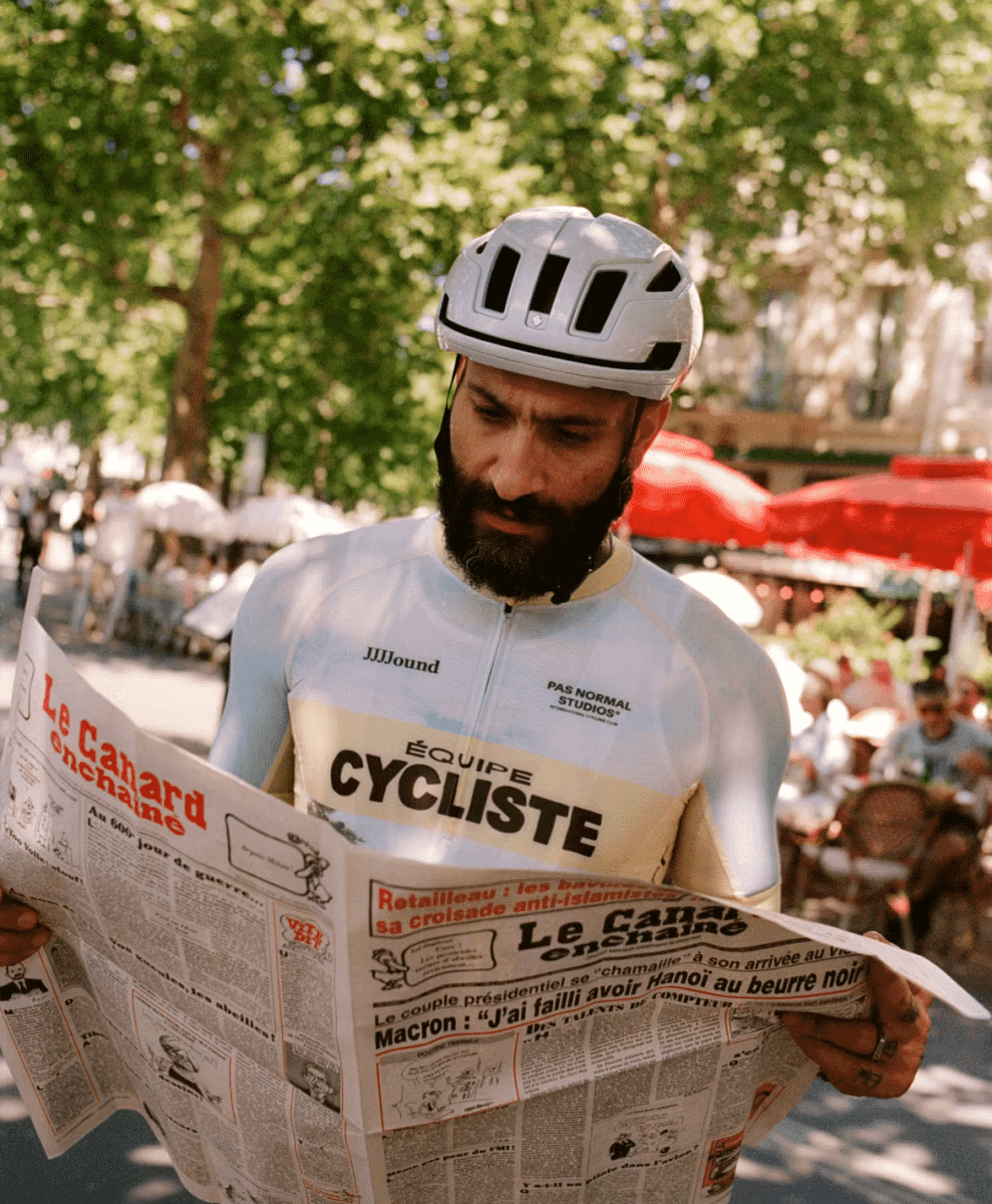 A man in a white cycling helmet and light blue jersey reading a newspaper outdoors.