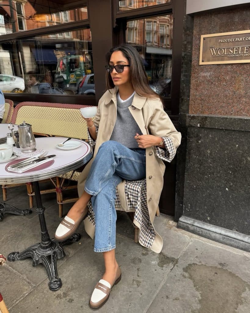 A woman wearing a beige trench coat, grey sweater, and blue jeans sitting at an outdoor café.