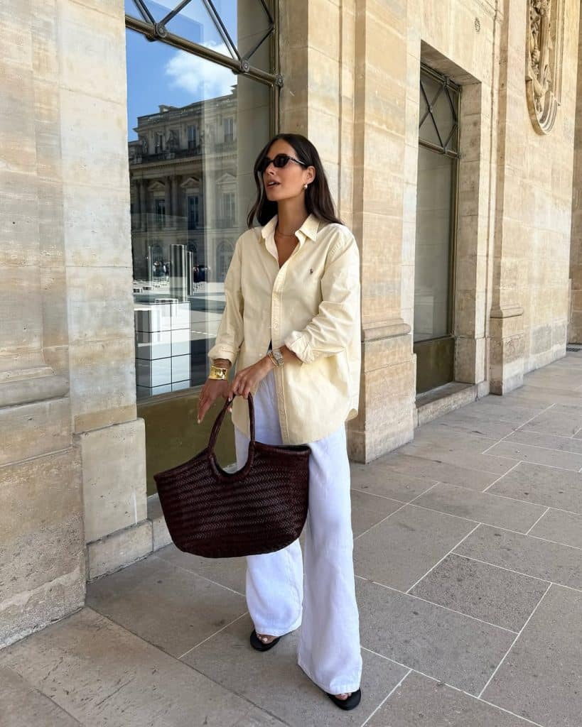A woman in an oversized yellow button-up shirt and white wide-leg pants carrying a woven tote.