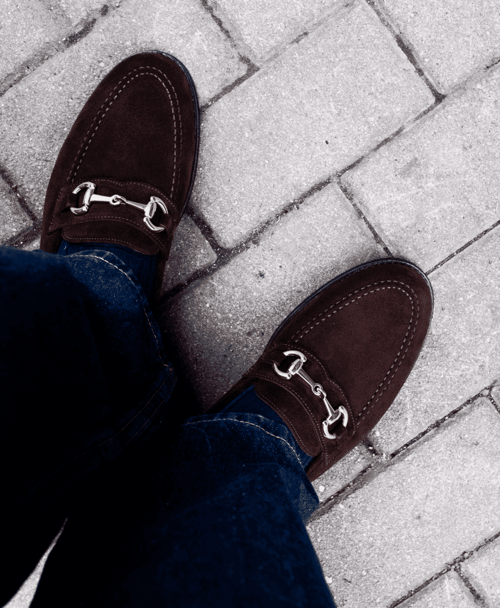 Overhead shot of brown suede loafers with silver horsebit buckles and denim jeans.