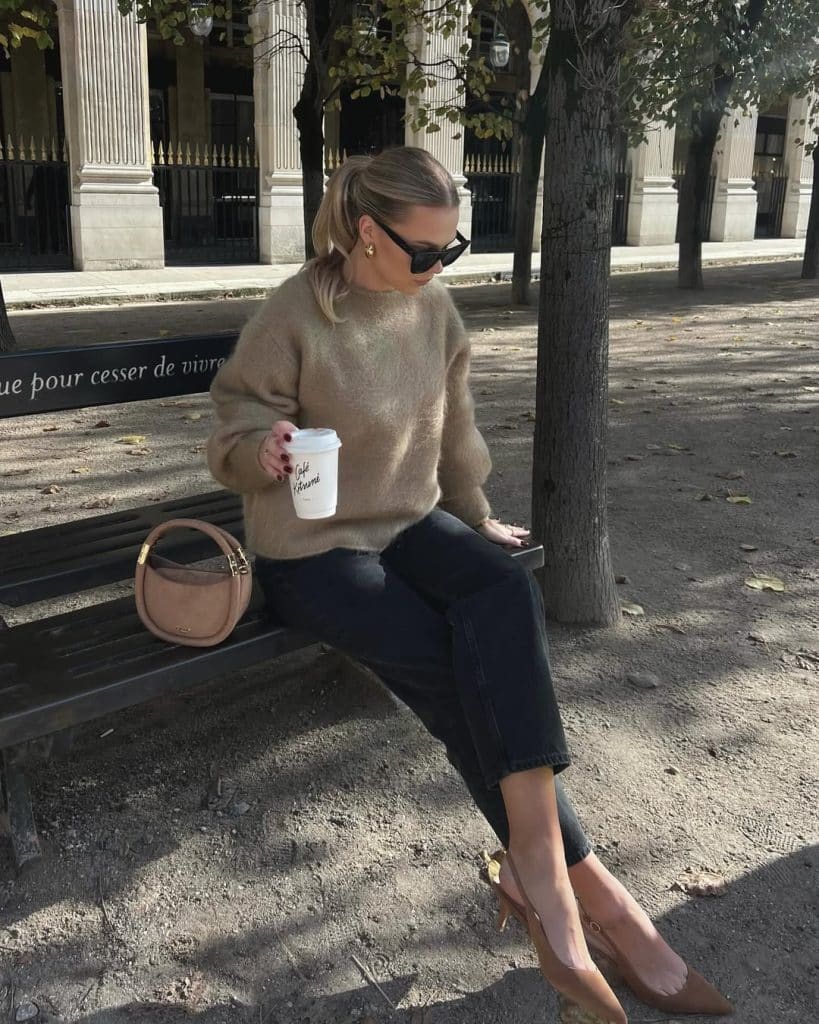 Woman in a beige fuzzy sweater and dark cropped jeans sitting on a park bench.