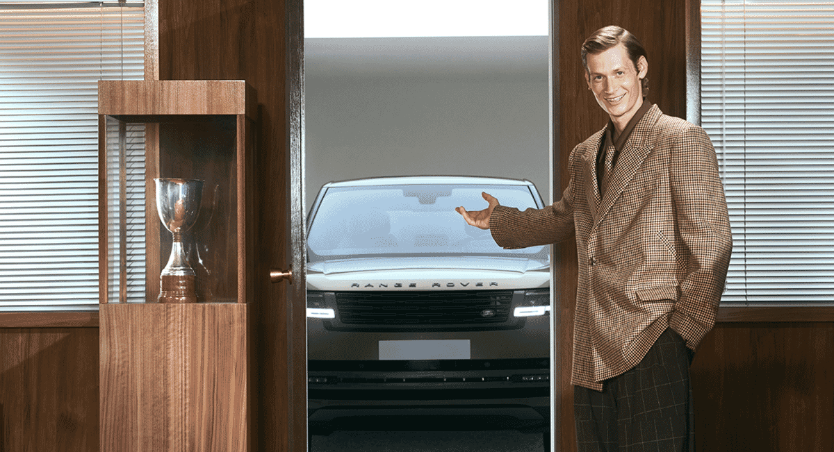 Man in a brown checkered wool coat gesturing toward a white Range Rover in a vintage setting.