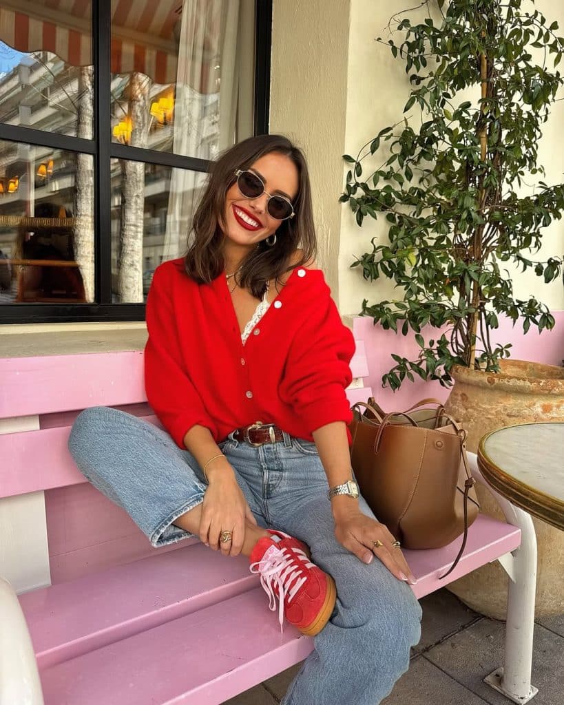 A woman in a bright red cardigan and light-wash jeans sitting on a pink bench at an outdoor café.