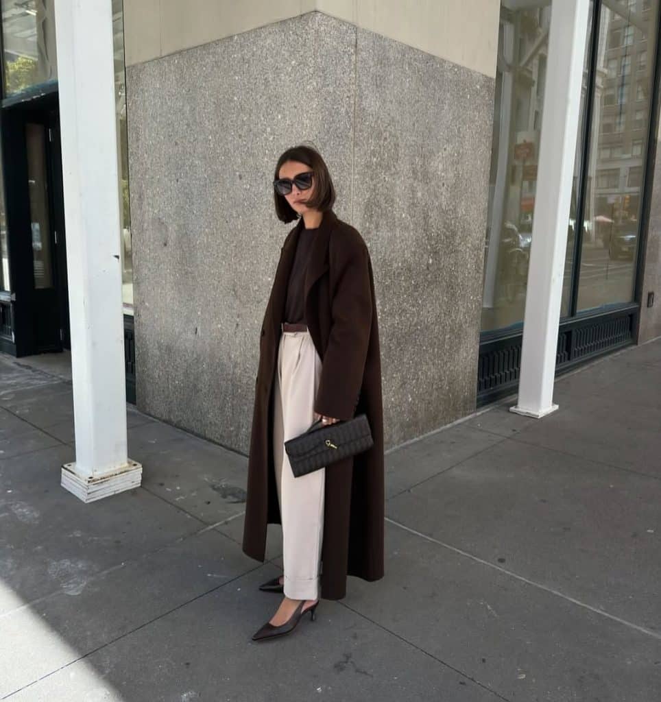 A woman in a long brown wool coat and cream wide-leg trousers walking on a sidewalk.