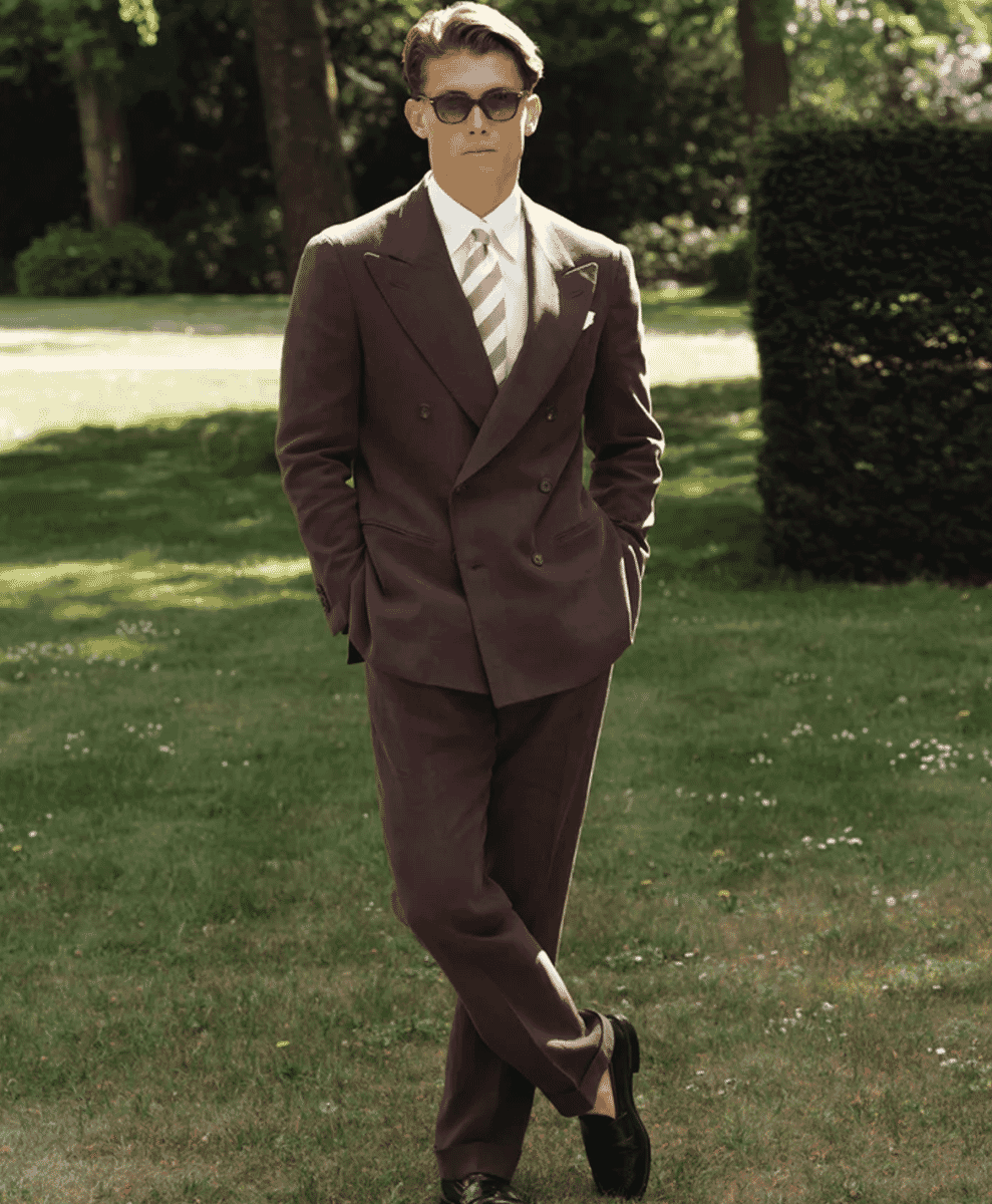 Man in a brown double-breasted suit walking on a sunlit lawn.