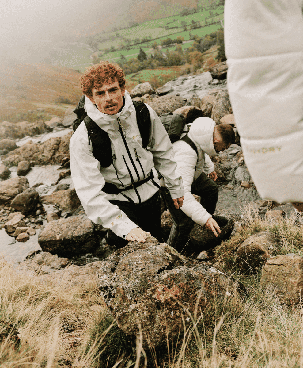 A man with curly hair wearing a white technical waterproof jacket while navigating a misty, rocky hillside.