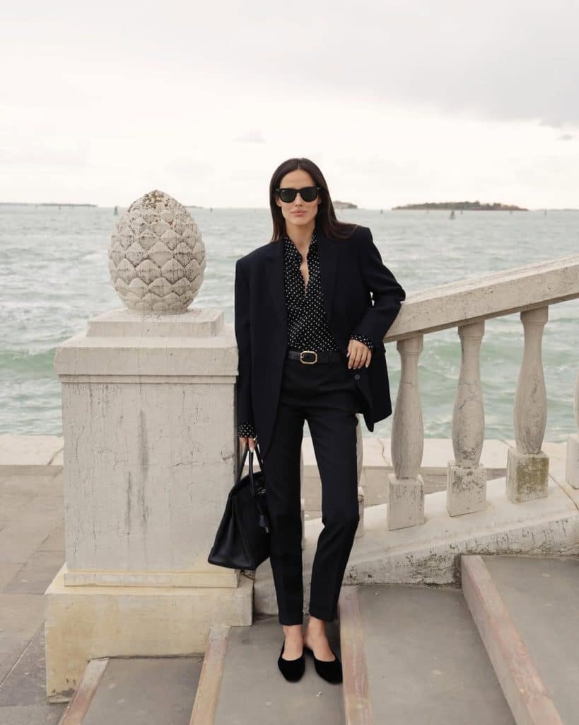A woman in an oversized black blazer and a polka-dot blouse standing by the water.