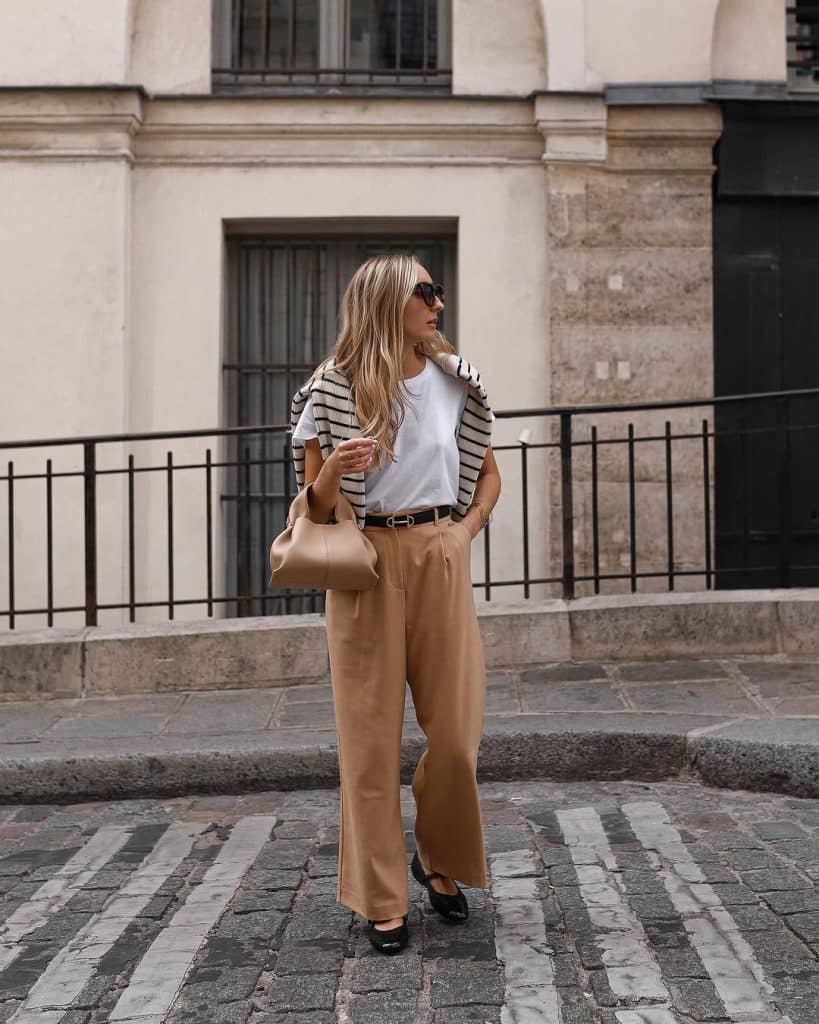 A woman in camel trousers and a striped cardigan walking on a cobblestone street.