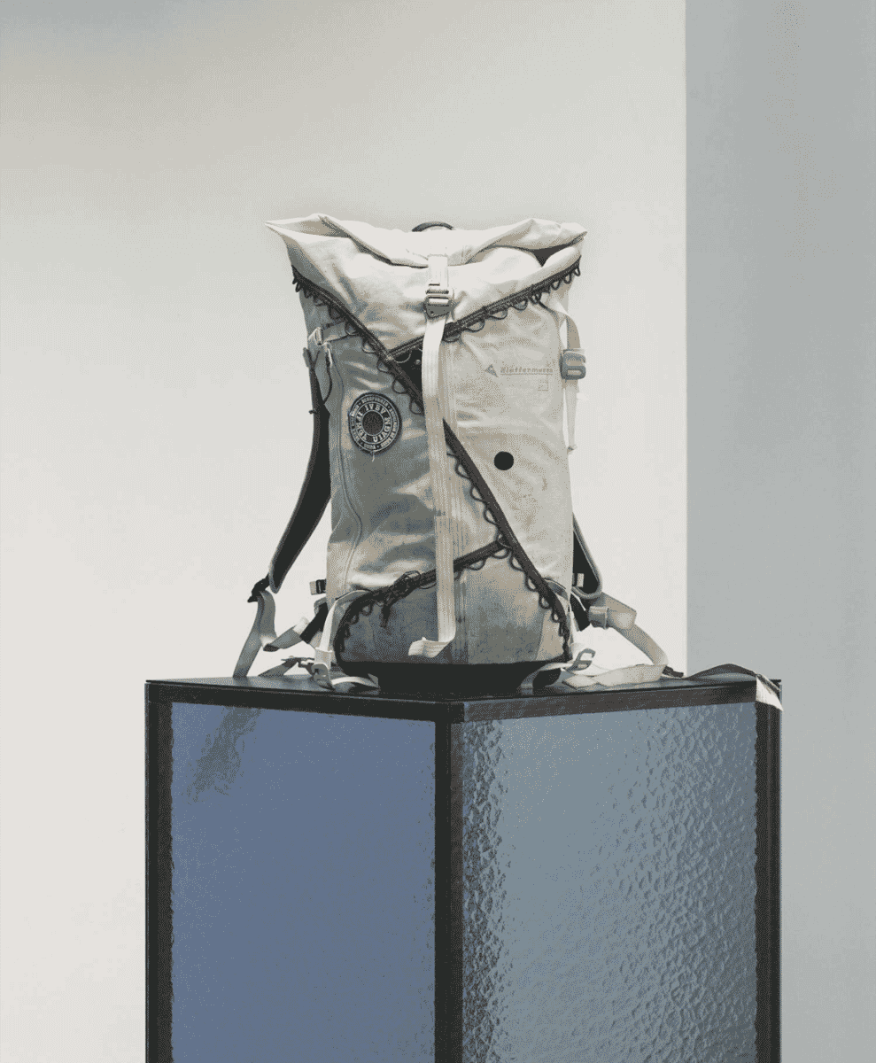A white Klättermusen backpack with detailed straps displayed on a stone pedestal.