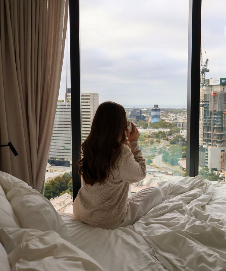 A woman in beige loungewear relaxes on a hotel bed with a mug while looking at the city skyline.