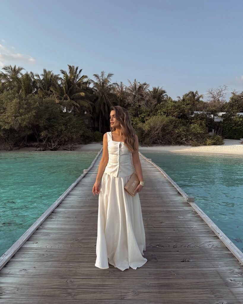 A woman in a white sleeveless corset-style top and flowing maxi skirt walking along a wooden pier.