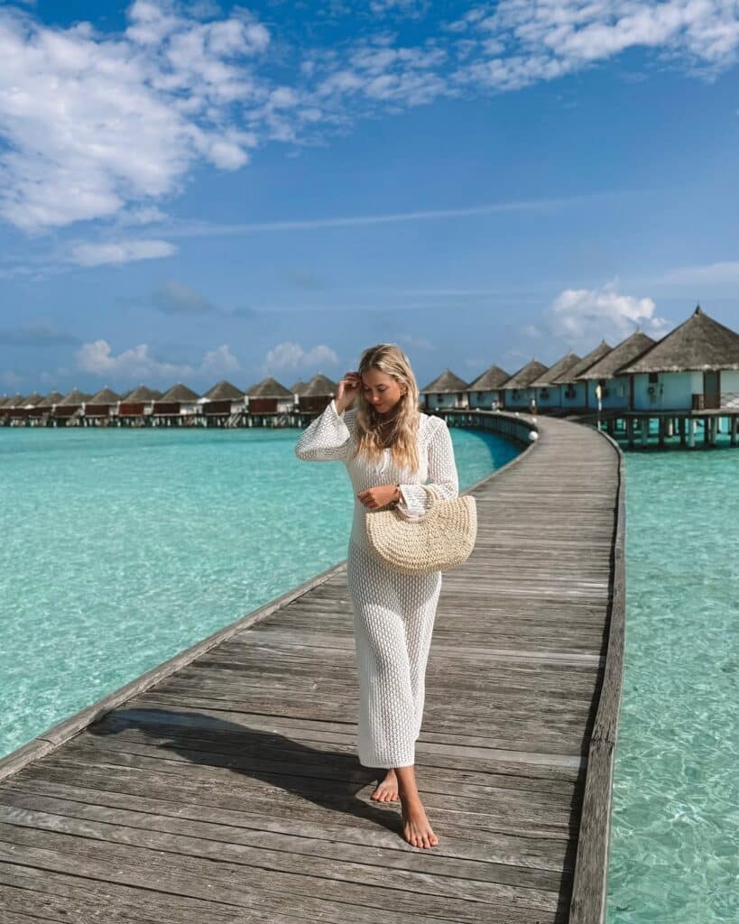 A brunette woman in a white knitted long-sleeve dress holding a straw bag on a wooden pier.