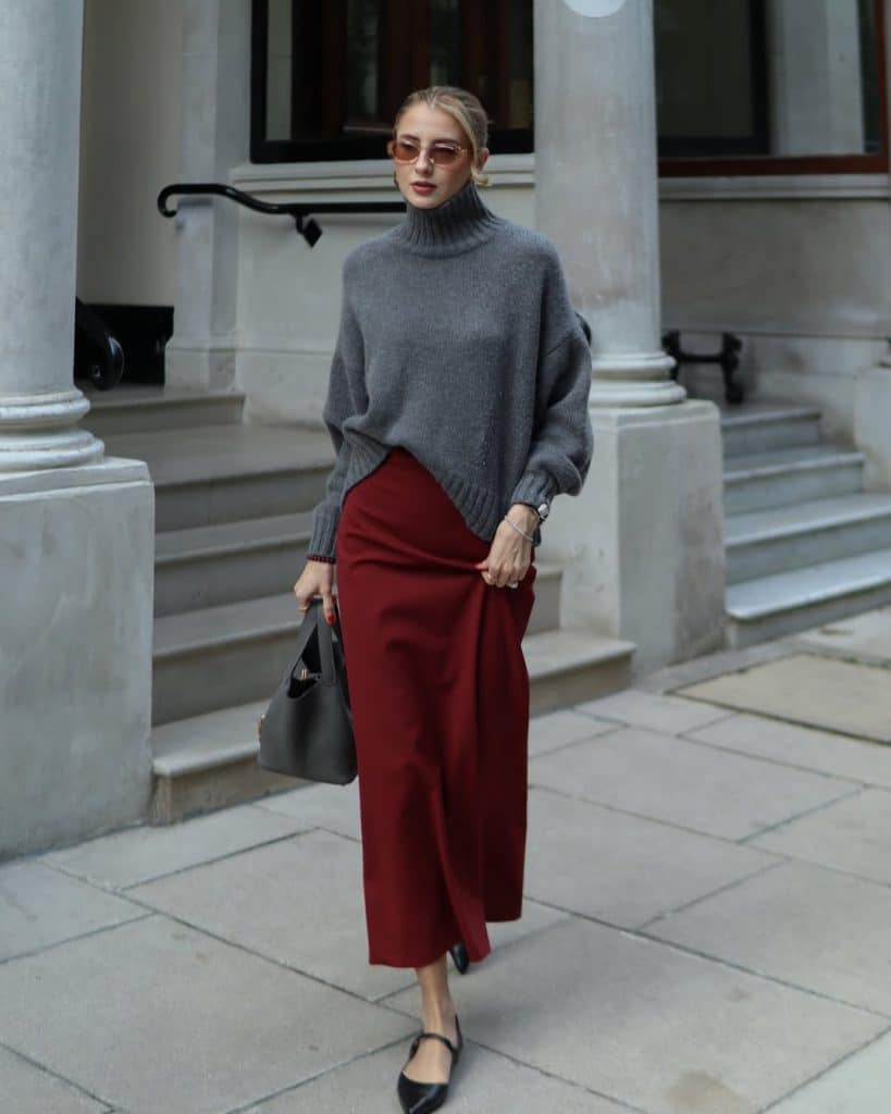Woman in an oversized grey turtleneck sweater and a flowing burgundy midi skirt walking on a sidewalk.