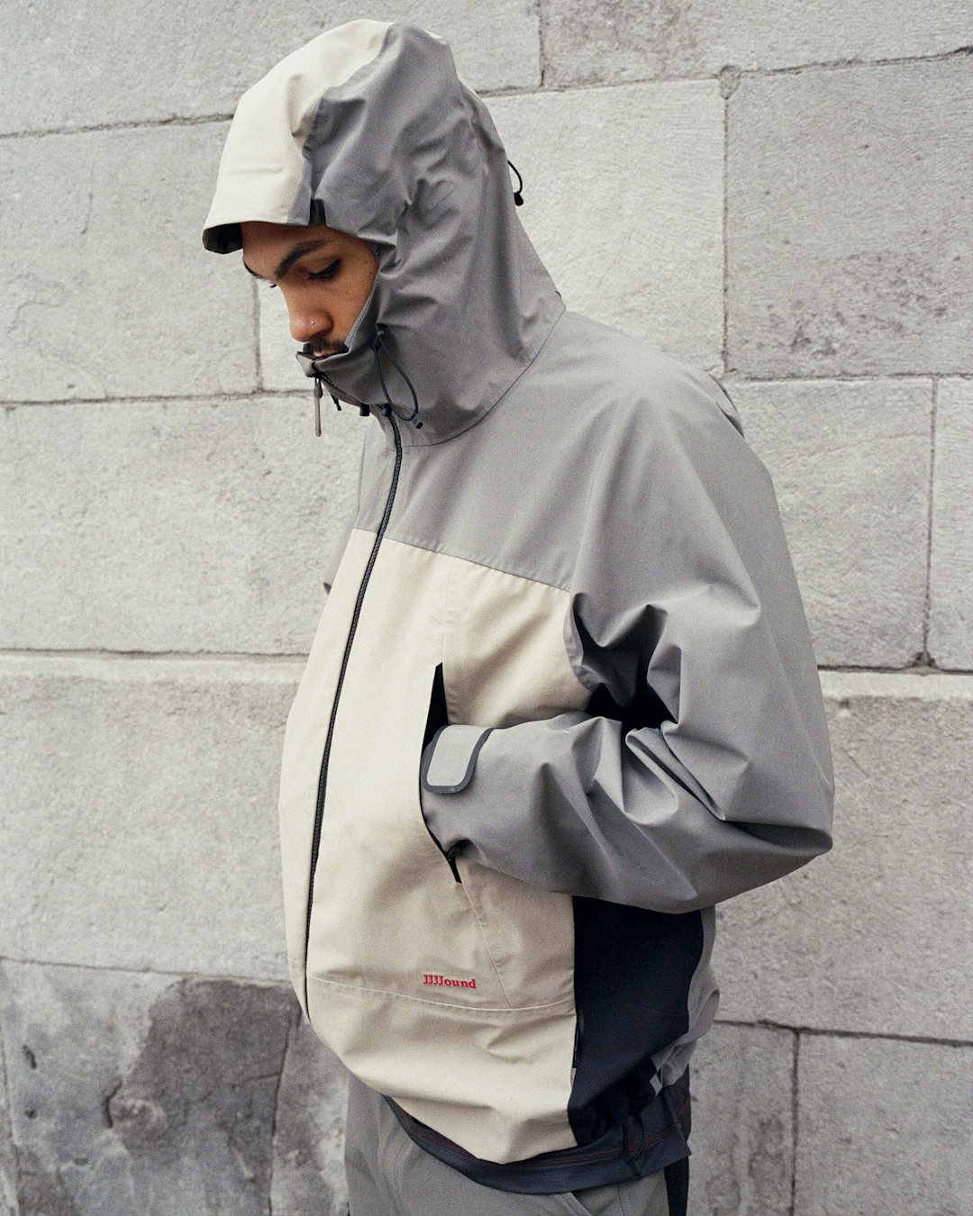 Close-up of a man in a color-blocked grey and beige hooded jacket with a red JJJJound logo.