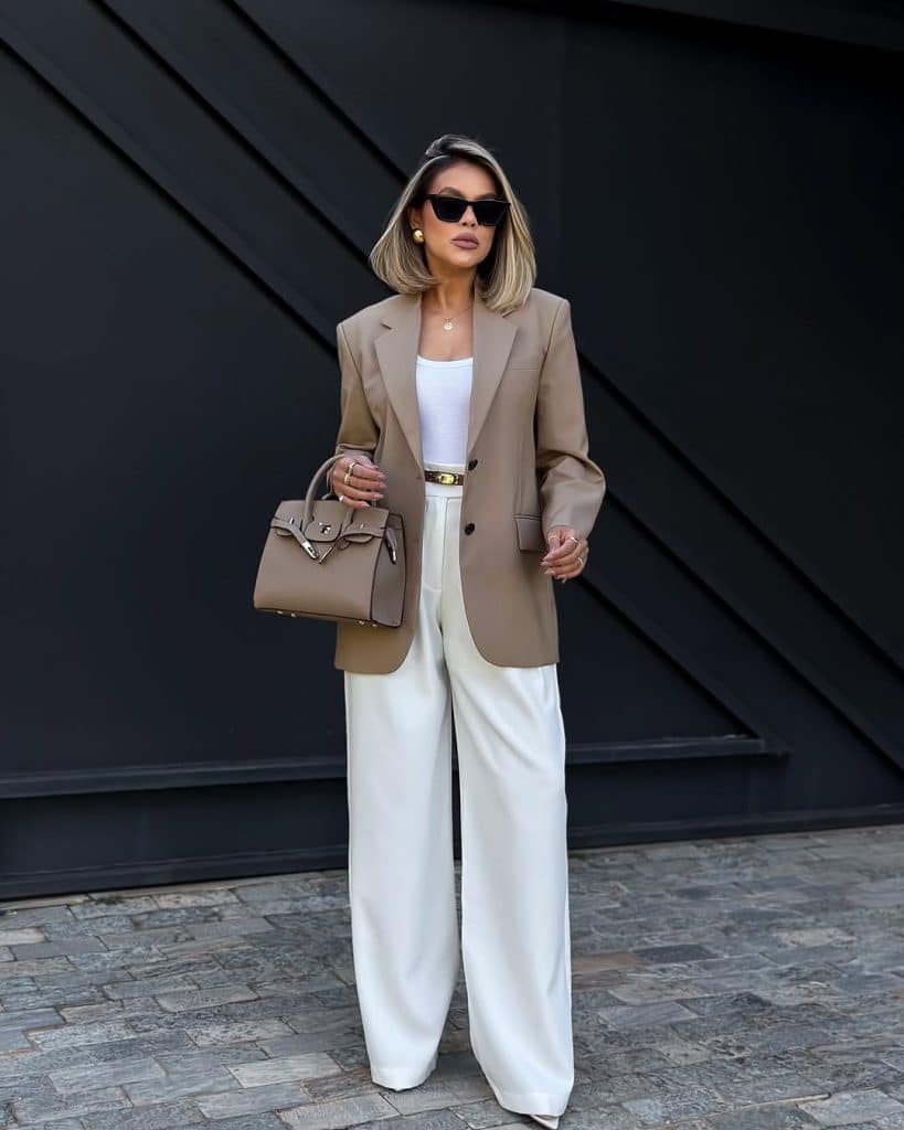 A woman in a beige blazer and white wide-leg trousers against a geometric wall.