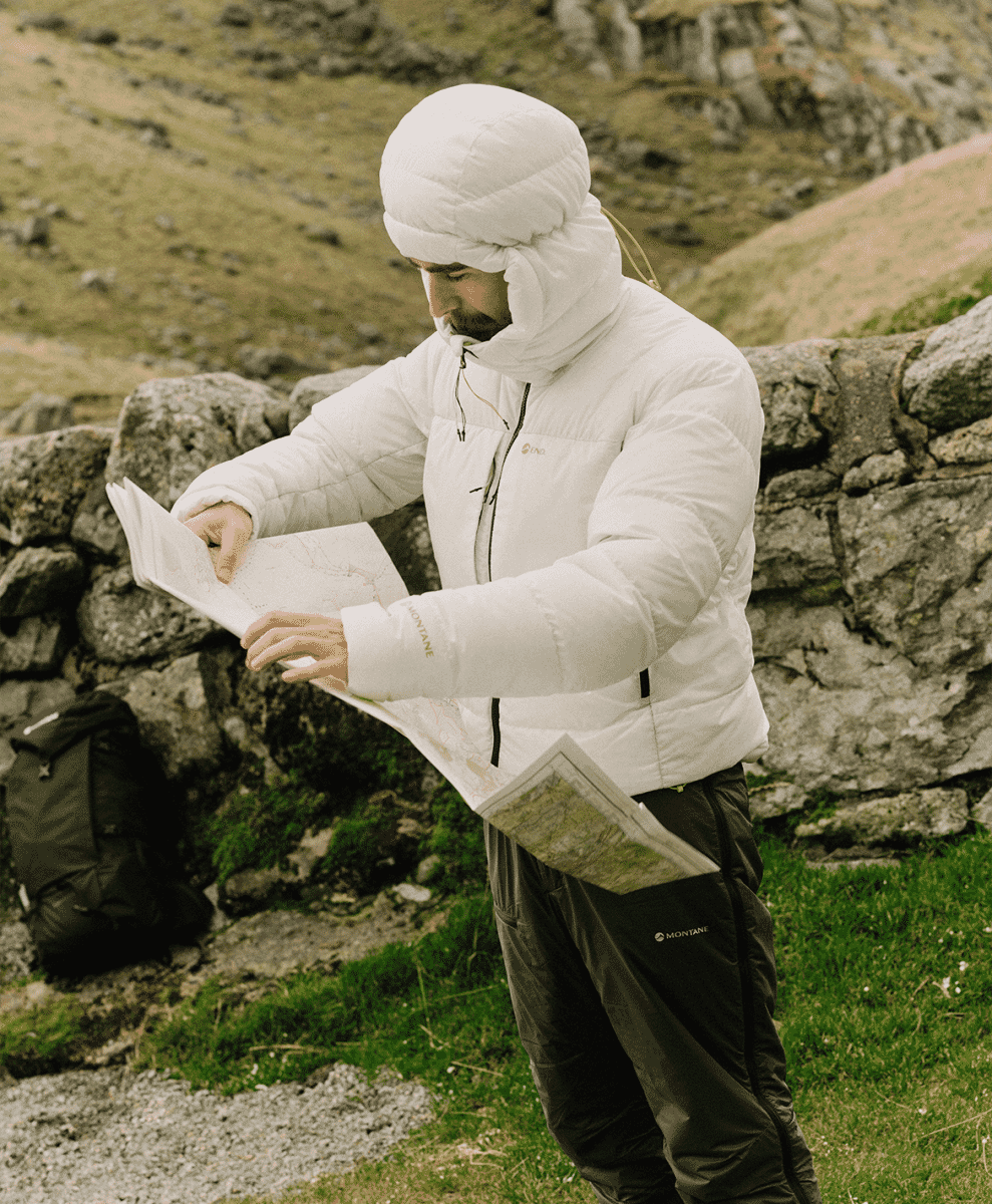 A hiker wearing a white puffer jacket and Montane branded pants consulting a map in a grassy, rocky field.