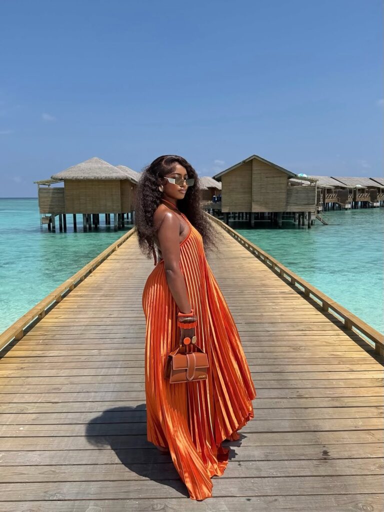 A woman in a vibrant orange pleated halter maxi dress on a wooden pier over turquoise water.