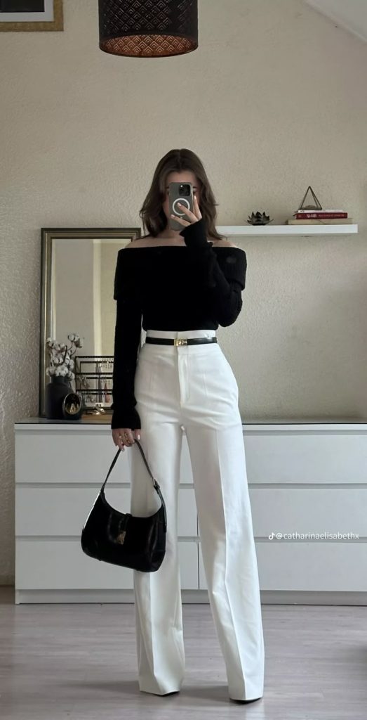 Mirror selfie of a woman wearing an off-the-shoulder black top and white wide-leg trousers with a gold belt.