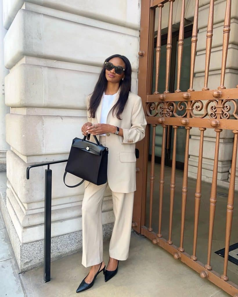 A woman in a tailored beige suit with black heels posing by a copper gate.