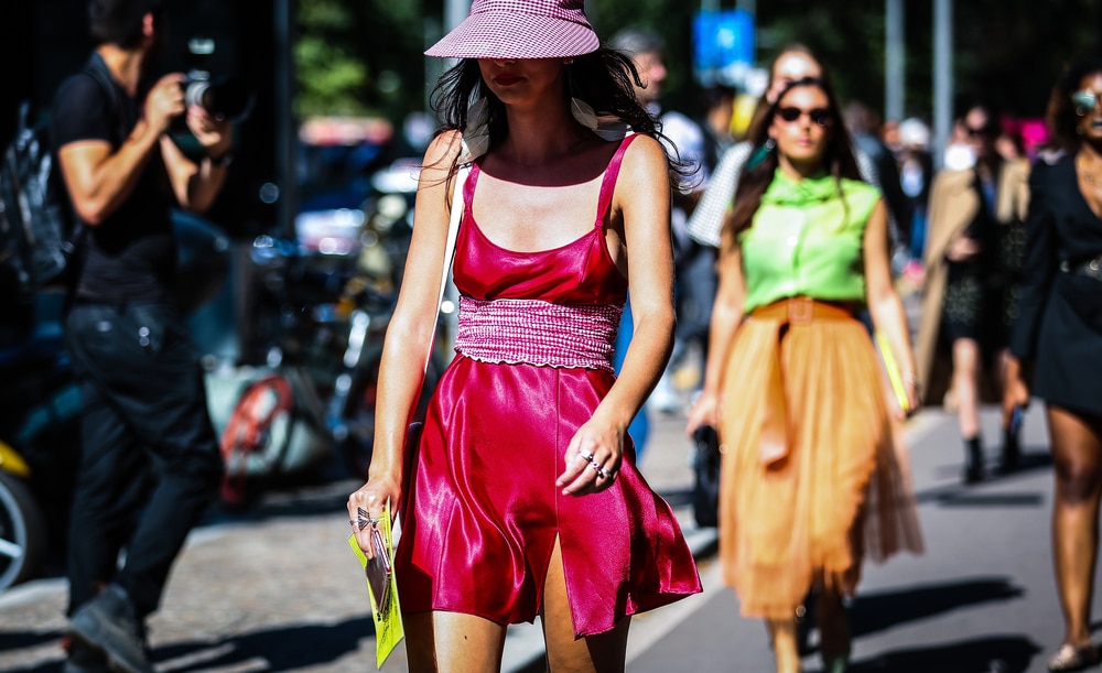 A woman in a vibrant red satin dress and gingham hat walking on a sunlit street.
