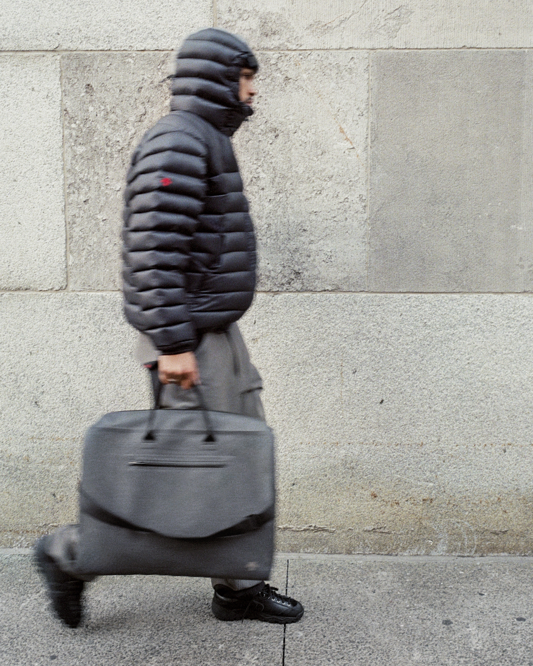 Motion blur shot of a man in a black puffer jacket carrying a large grey duffel bag.