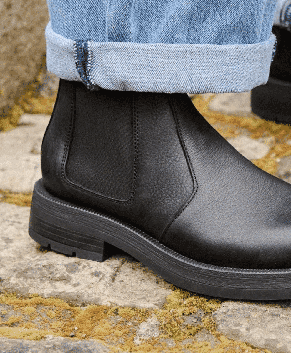 Black leather Chelsea boots worn with light-wash denim on mossy stone pavement.