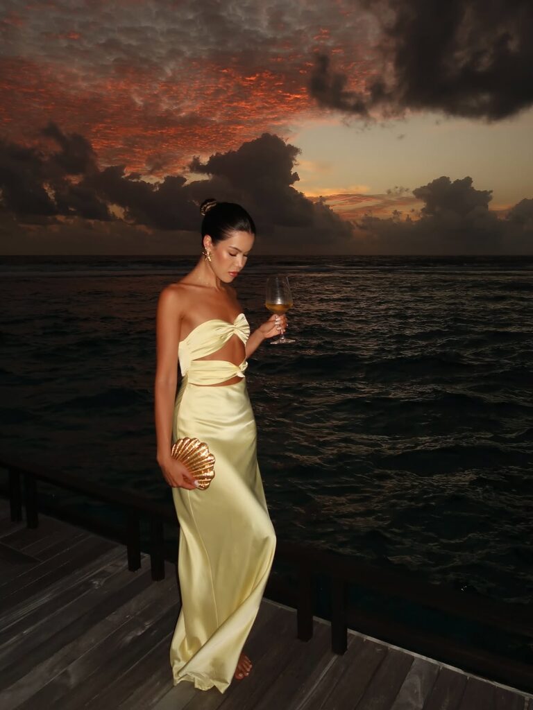 A woman in a pale yellow satin gown holding a shell clutch on a wooden pier at sunset.